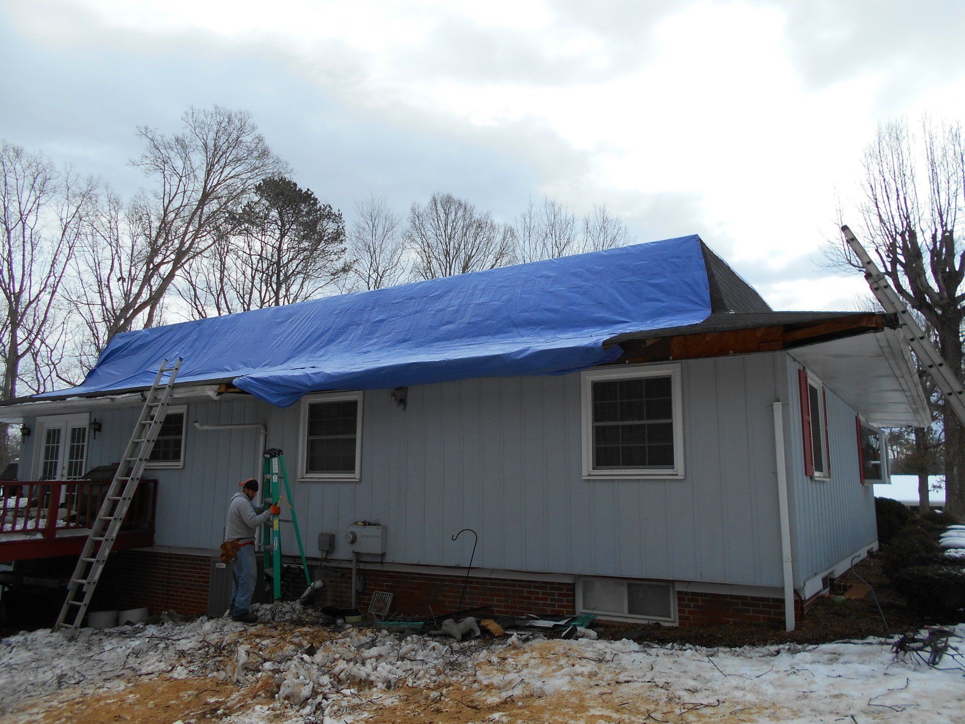 A blue tarp covers a damaged house roof. Two men work on the roof repair. Snow on the ground.