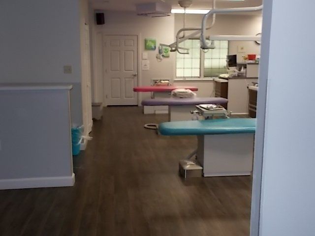 Dentist's office with three examination tables. Teal, pink, and white tables on wood floor, with windows in the background.