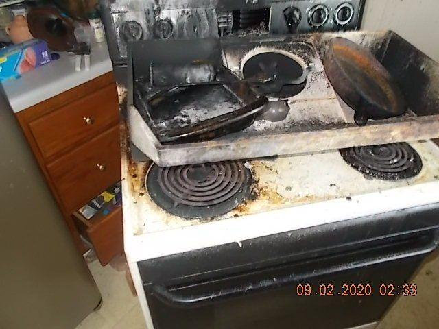 Damaged stovetop with burnt pans and residue from a fire. Cabinet and refrigerator in the background.