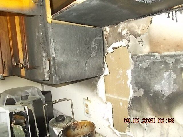 Charred kitchen interior showing damaged cabinets, appliances, and wall with a timestamp.