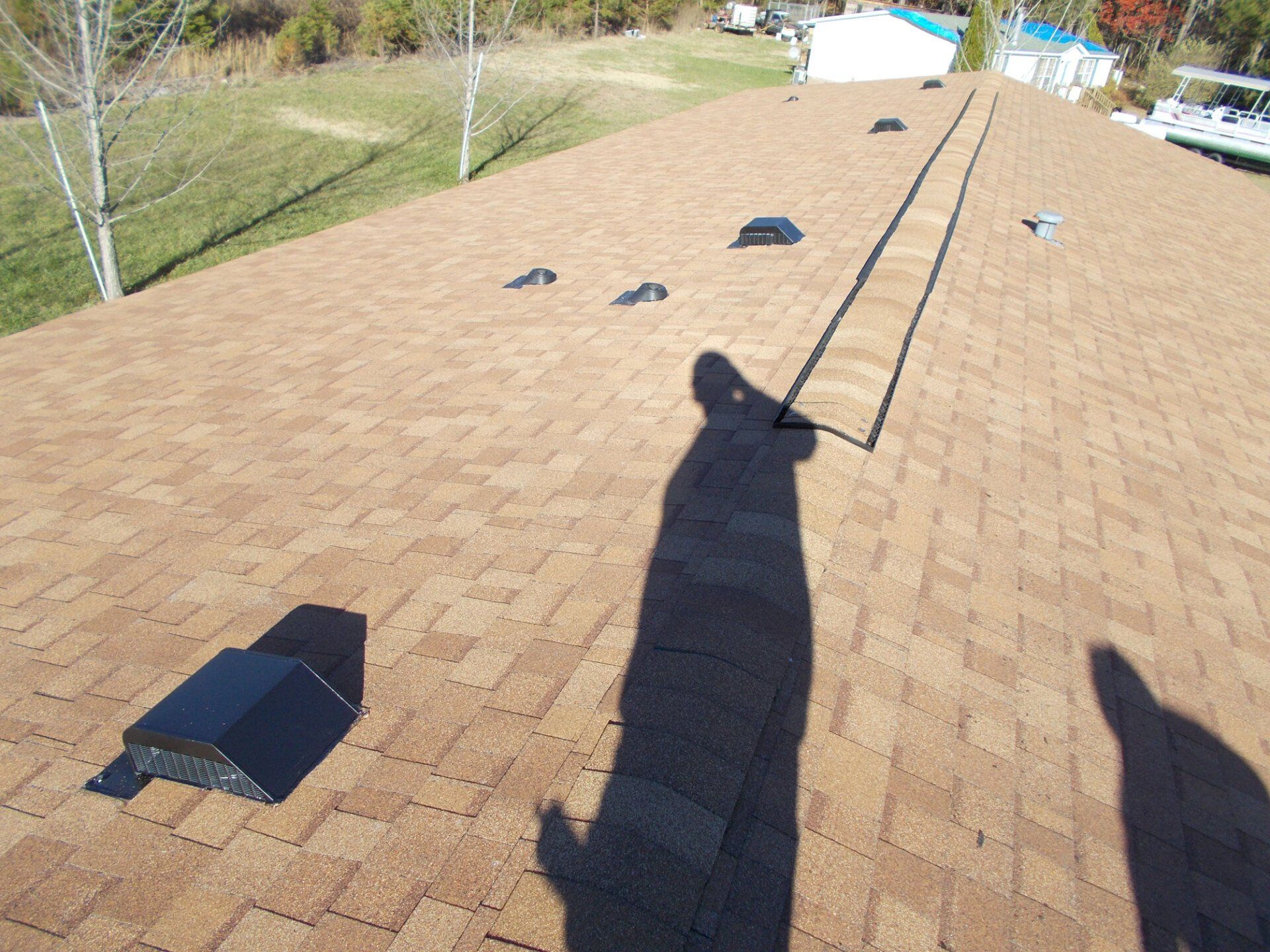 Brown shingled roof with black vents, person's shadow cast across it on a sunny day.
