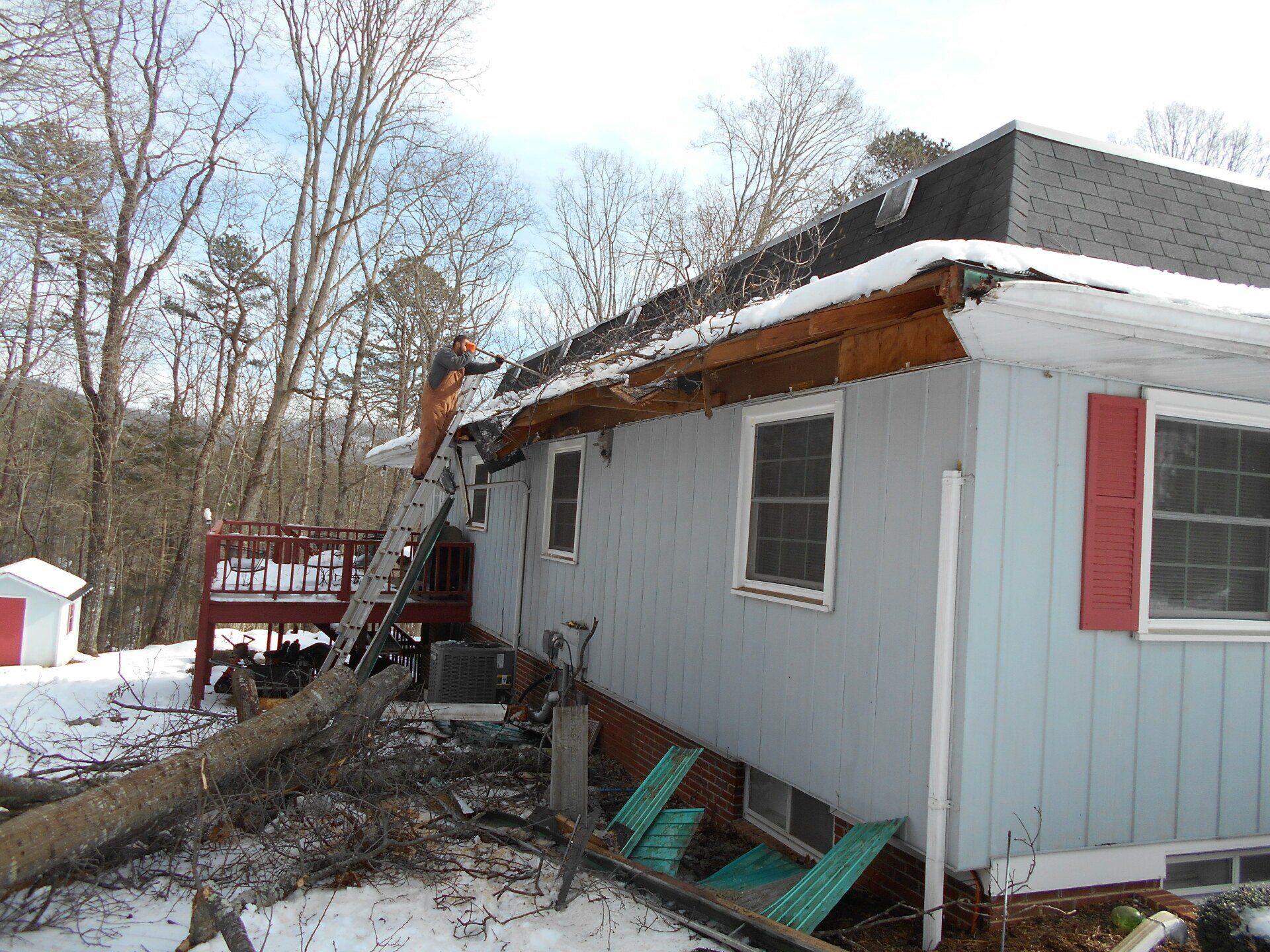 Person on ladder repairing house roof with snow. Light blue siding, red shutters, winter setting.