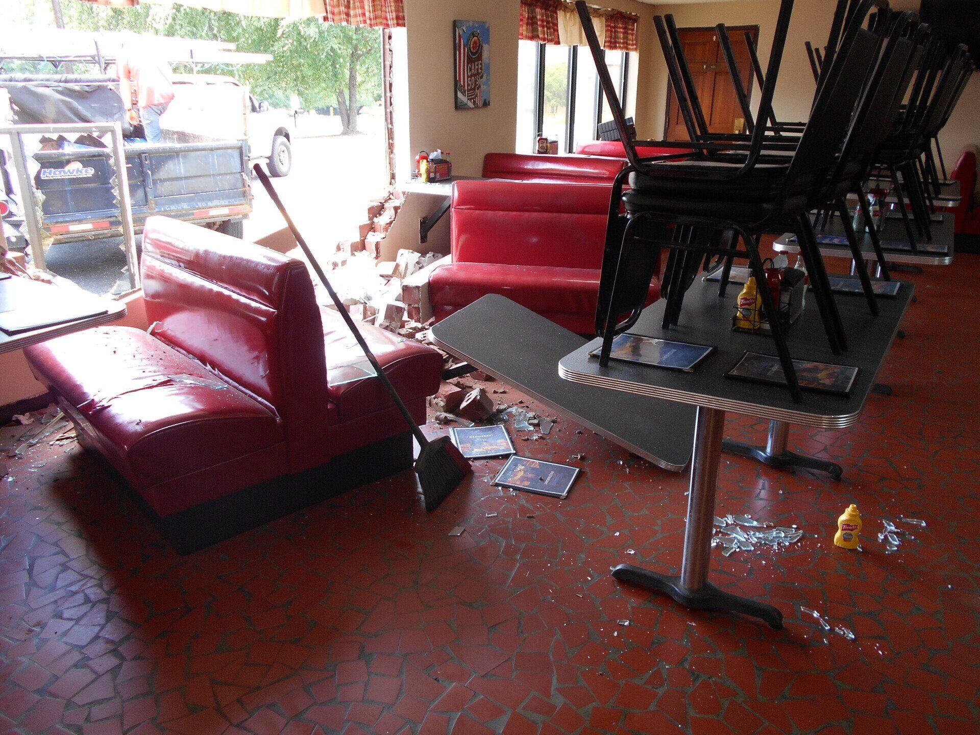 Interior of a diner wrecked, red booth, tables with chairs stacked on them, broken glass on the floor, daylight streaming through the window.