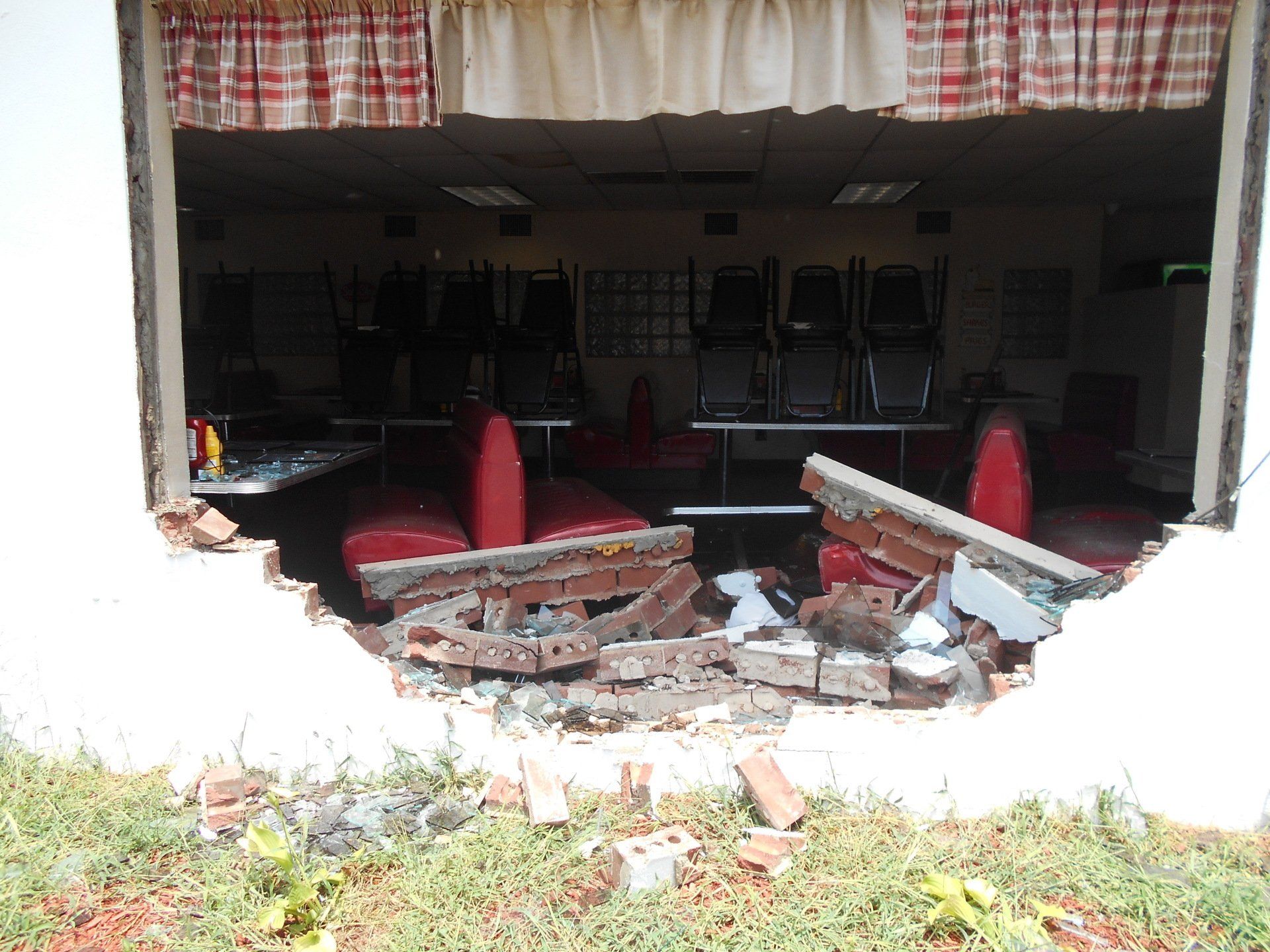 A brick wall with a large hole reveals an interior space with chairs and red seating; daylight visible.