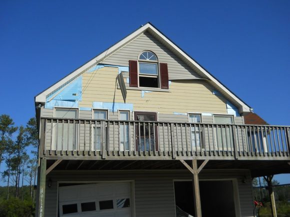 A dilapidated two-story house with a deck, detached siding, and broken garage door against a clear blue sky.