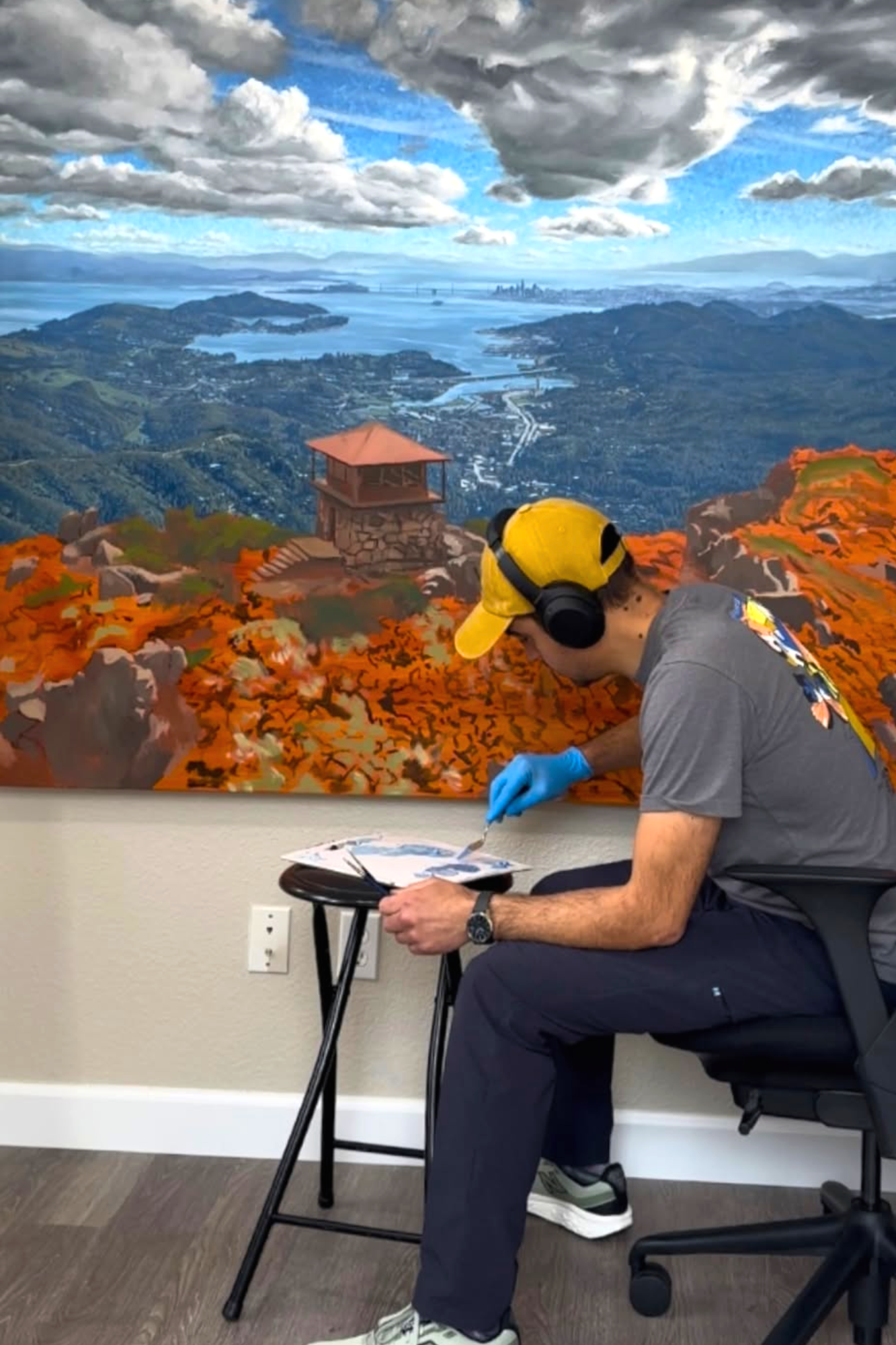 Person in yellow cap painting a mountain landscape mural on a wall in a room