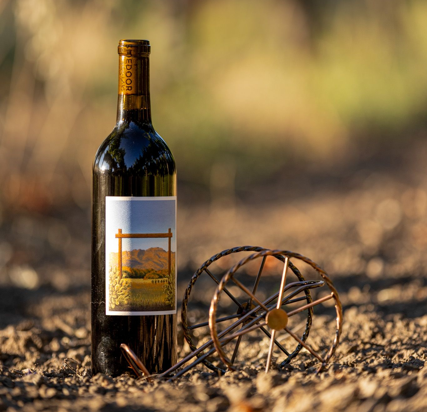 Wine bottle beside a rustic wagon wheel on dry ground, with a blurred field in the background