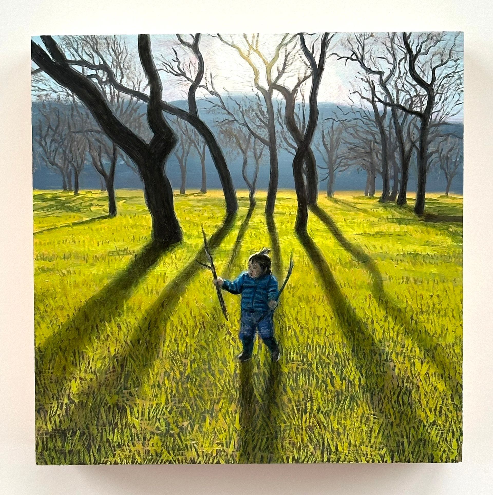 Person in blue stands in a sunlit grove of leafless trees casting long shadows over green grass.