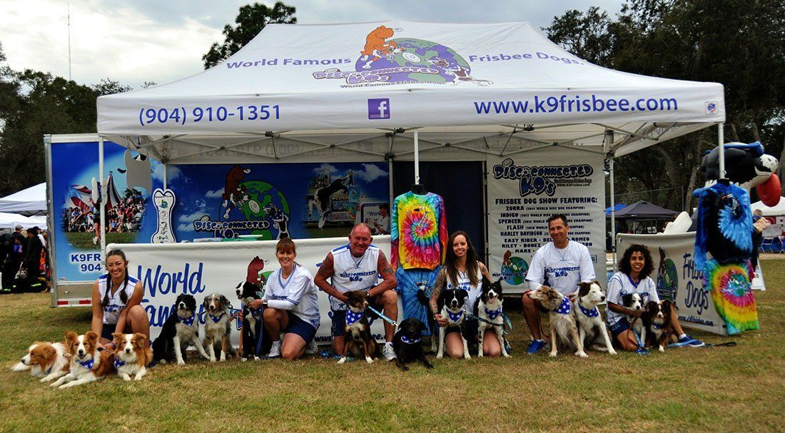 Group posing under a white event tent with colorful artwork and banners on a grassy field