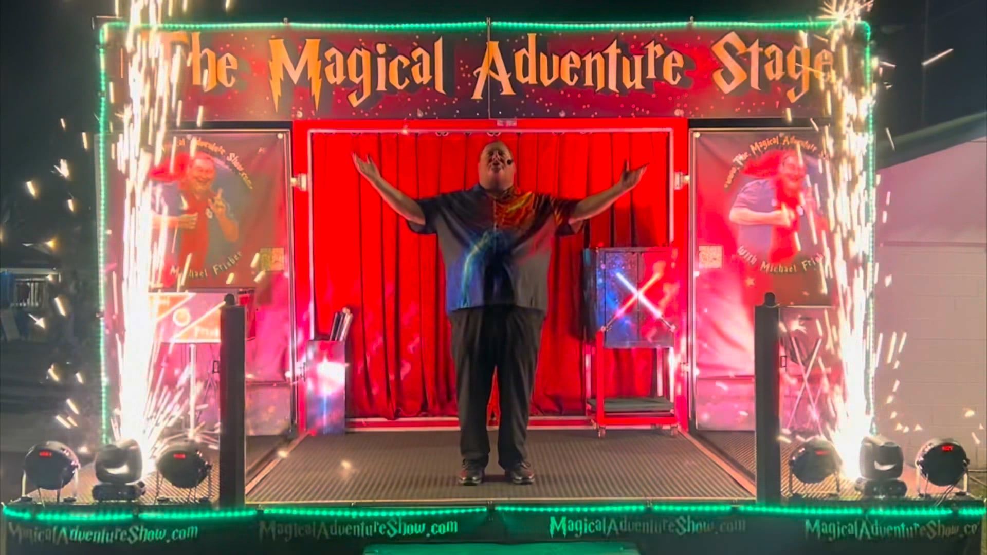 Performer stands center stage with arms outstretched before red curtains and colorful lights at The Magical Adventure Stage