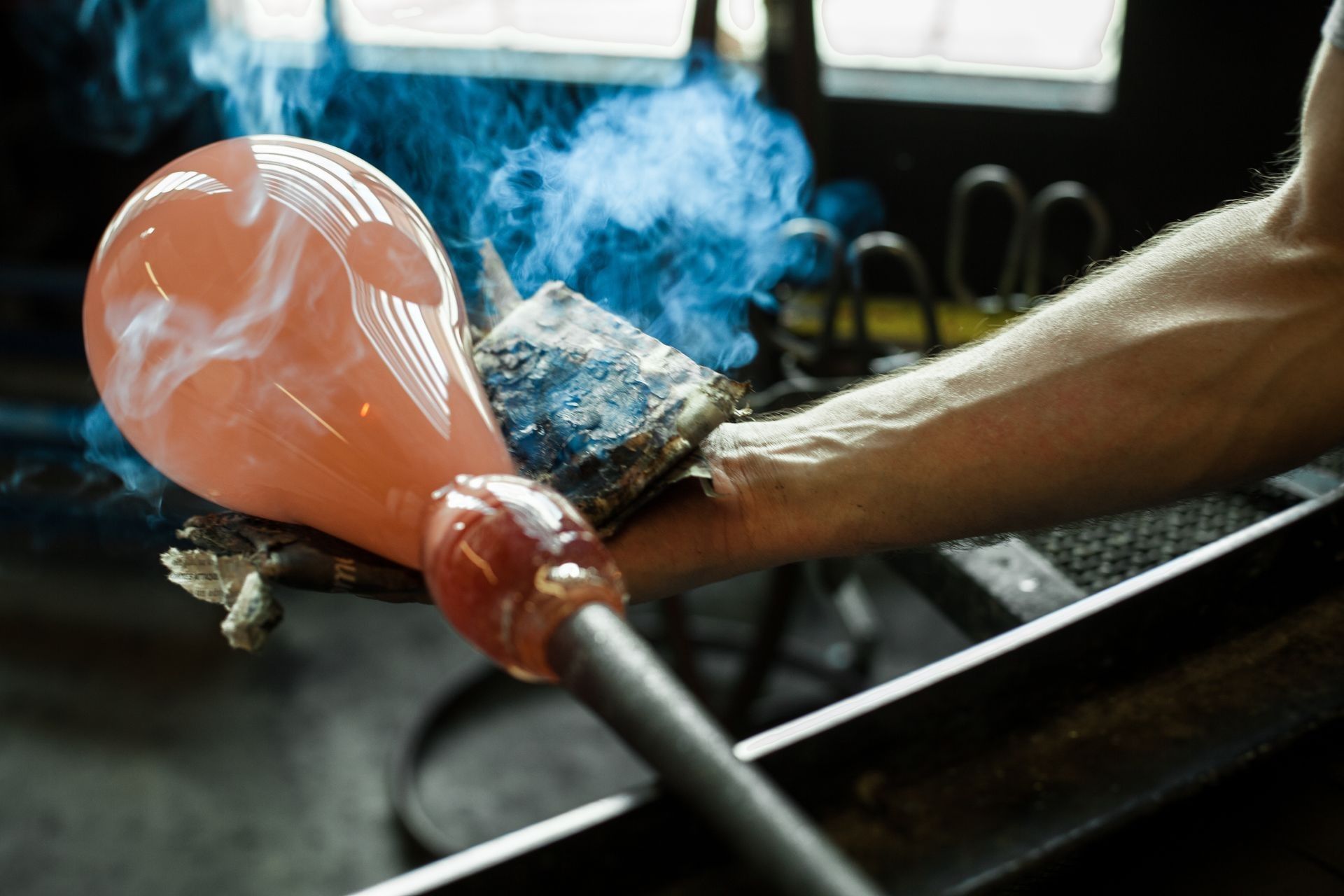 Molten orange glass being shaped with metal tools, with blue smoke in a workshop sink