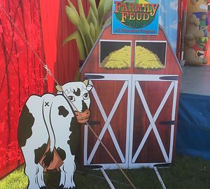 Cow cutout beside a red barn-style Family Feud game display with yellow pieces and green backdrop