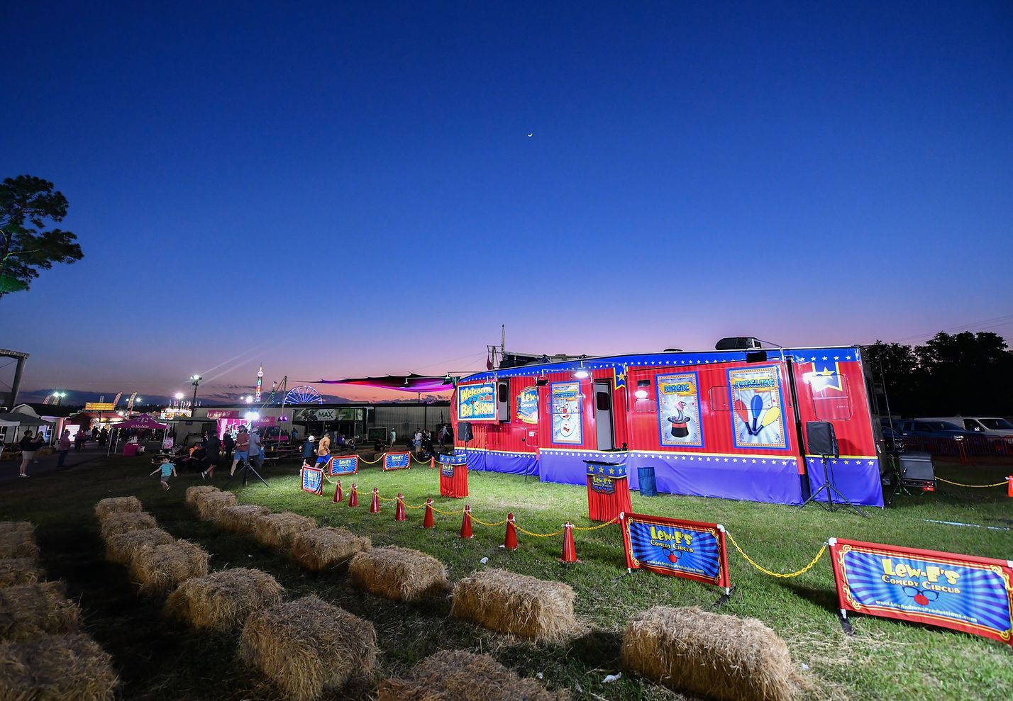 Colorful fairground tent at dusk with hay bales, flags, and people under a blue evening sky