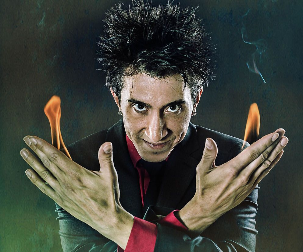 Dark-haired person in a black and red outfit, crossing arms with flames rising from both hands against a dark background