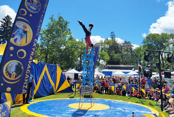 Acrobat balancing atop a ladder on a blue mat at an outdoor circus festival with a crowd watching