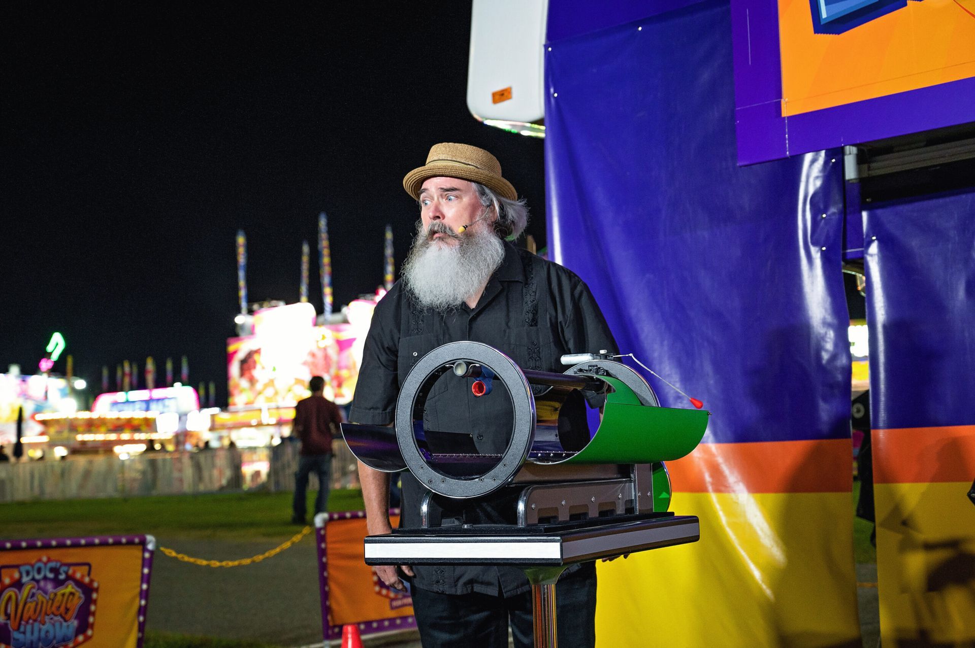 Bearded man in a straw hat beside a machine at a nighttime carnival, with colorful tents in the background