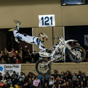 Motorcyclist performing a stunt jump indoors beneath sign 121, with spectators watching.