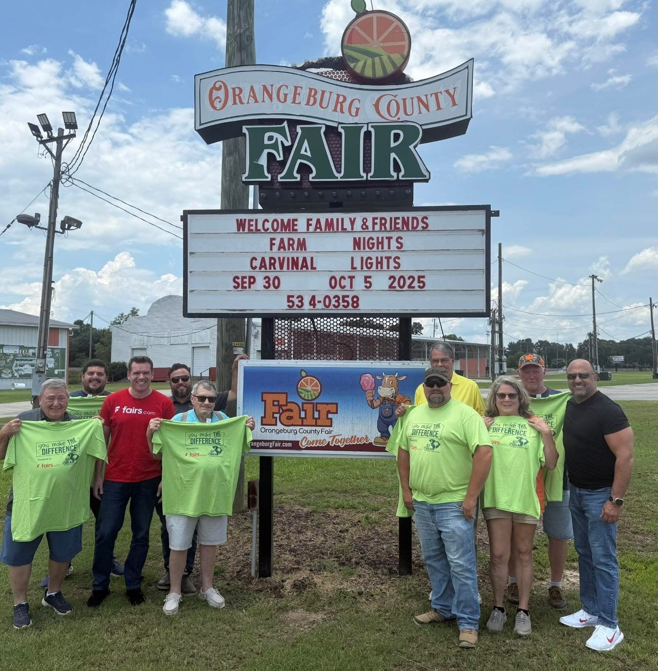 Group posing by Orangeburg County Fair sign under a cloudy sky, with neon shirts and fair tent info displayed.