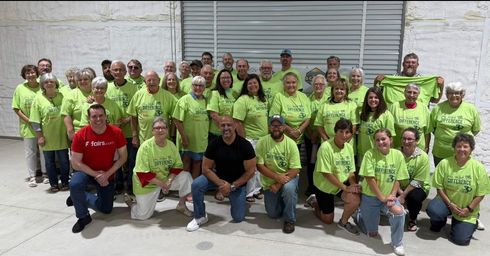 Group of volunteers in neon green shirts posing in front of a white building with a red-shirted man kneeling in front
