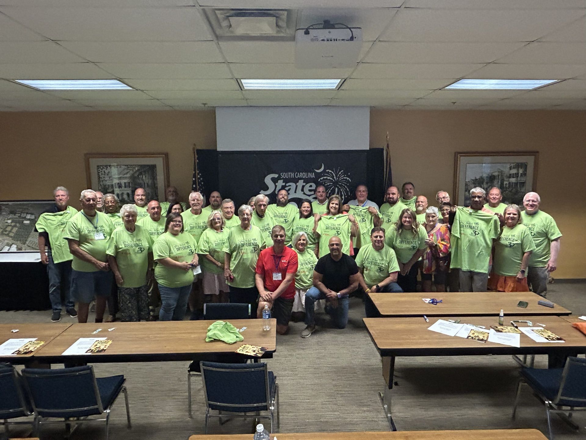 Group of volunteers in matching green shirts posing indoors behind tables at a community event