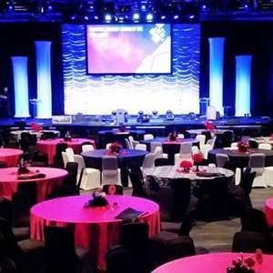 Conference banquet hall with round tables, pink tablecloths, and a lit stage with a large screen