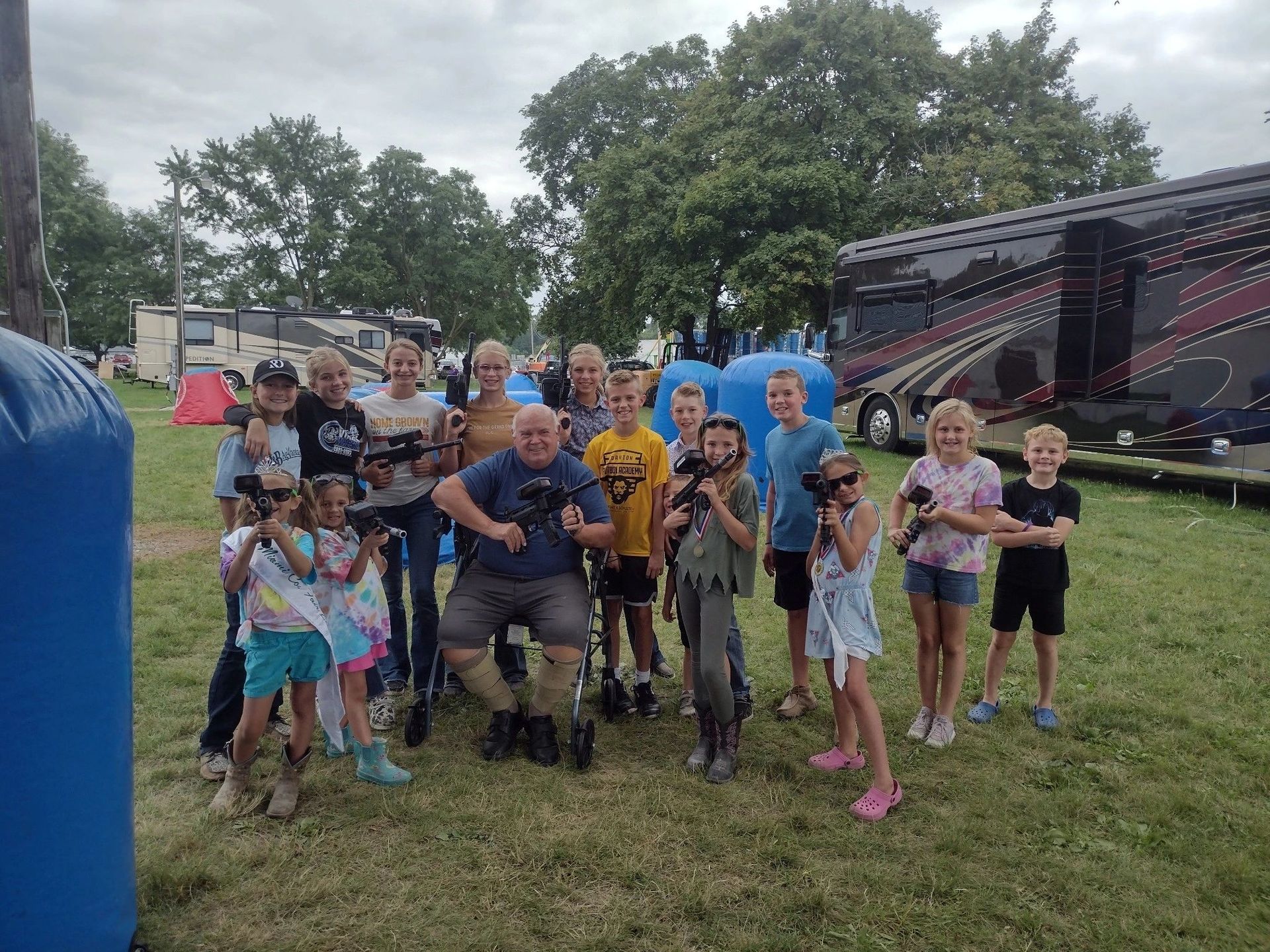 Group posing outdoors on a grassy field beside a large RV, with two people in blue costumes in back.