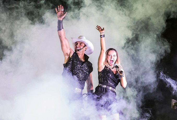 Two performers raise their arms in thick white stage smoke, wearing black outfits and hats.