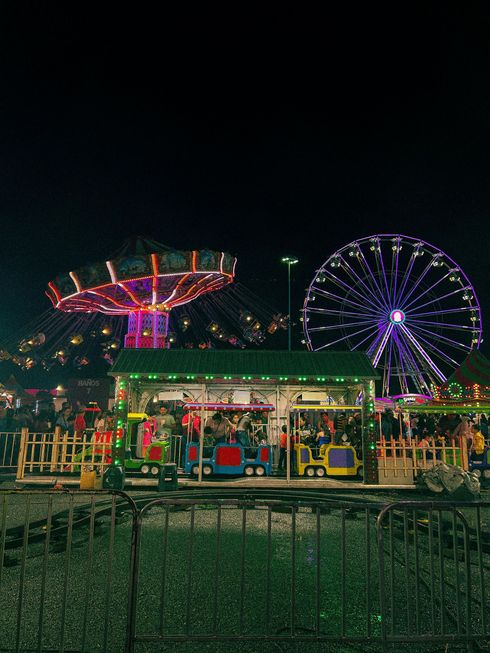 Nighttime carnival with a brightly lit swing ride, Ferris wheel, and colorful booths behind a fence.