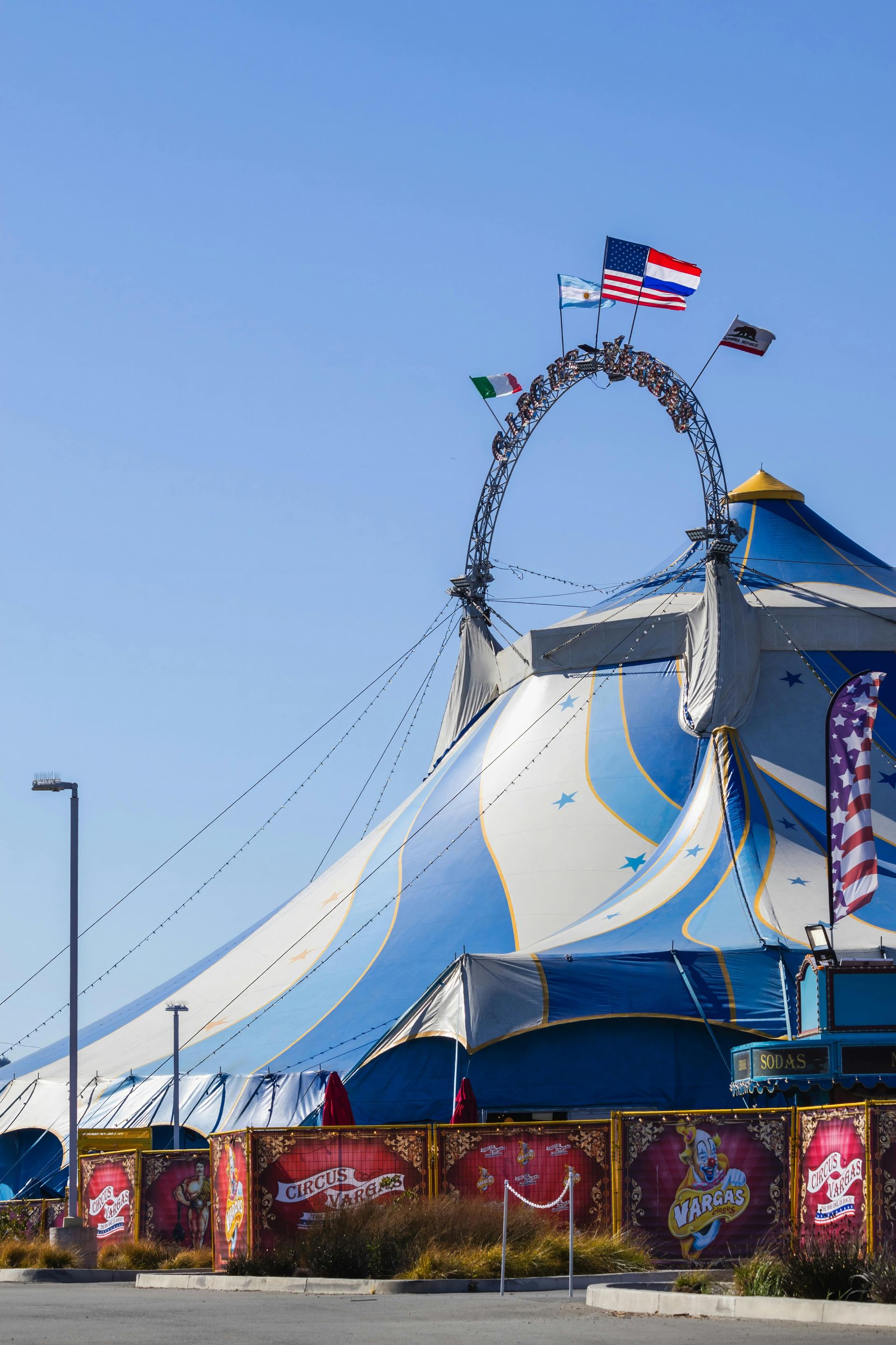 Blue-and-white circus tent with a tall hoop and flags against a clear sky