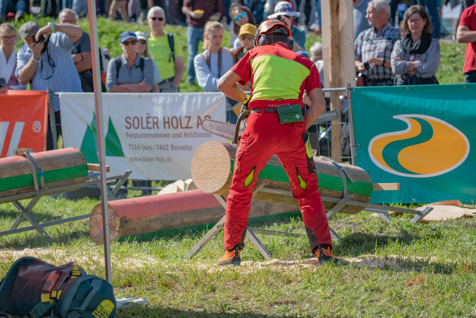 Red-clad lumberjack chopping a log at an outdoor competition, with spectators behind barriers.