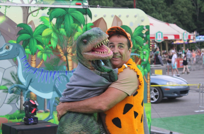 Person in a dinosaur costume hugging a smiling person in a Flintstones-style outfit at an outdoor event