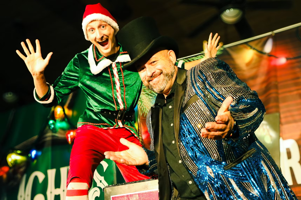 Two performers onstage, one in green and red, one in a blue jacket and black hat, both posing energetically