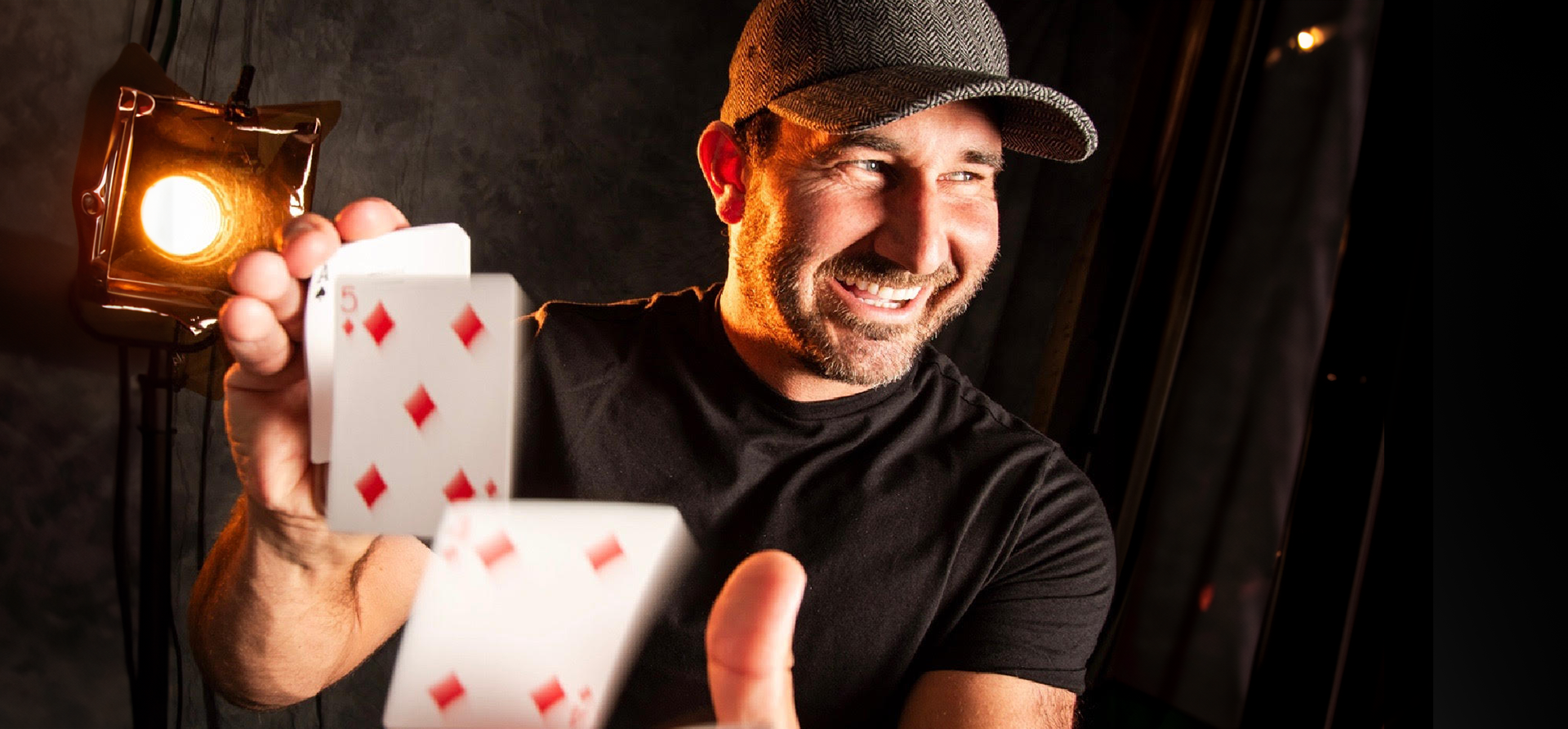 Man holding playing cards and smiling beside a glowing lantern in a dark setting