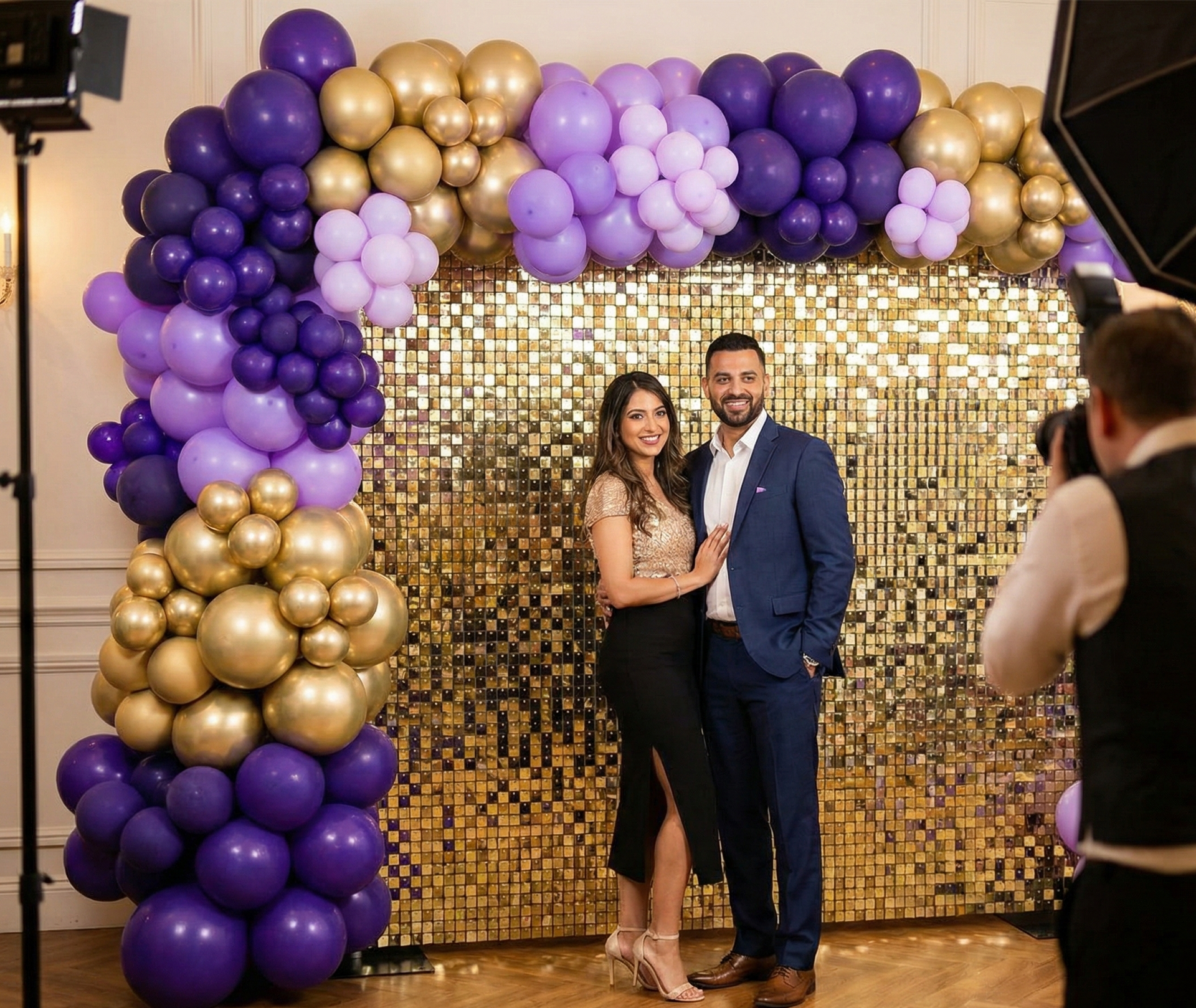 Couple posing in front of a gold sequin wall backdrop with purple and gold balloon styling