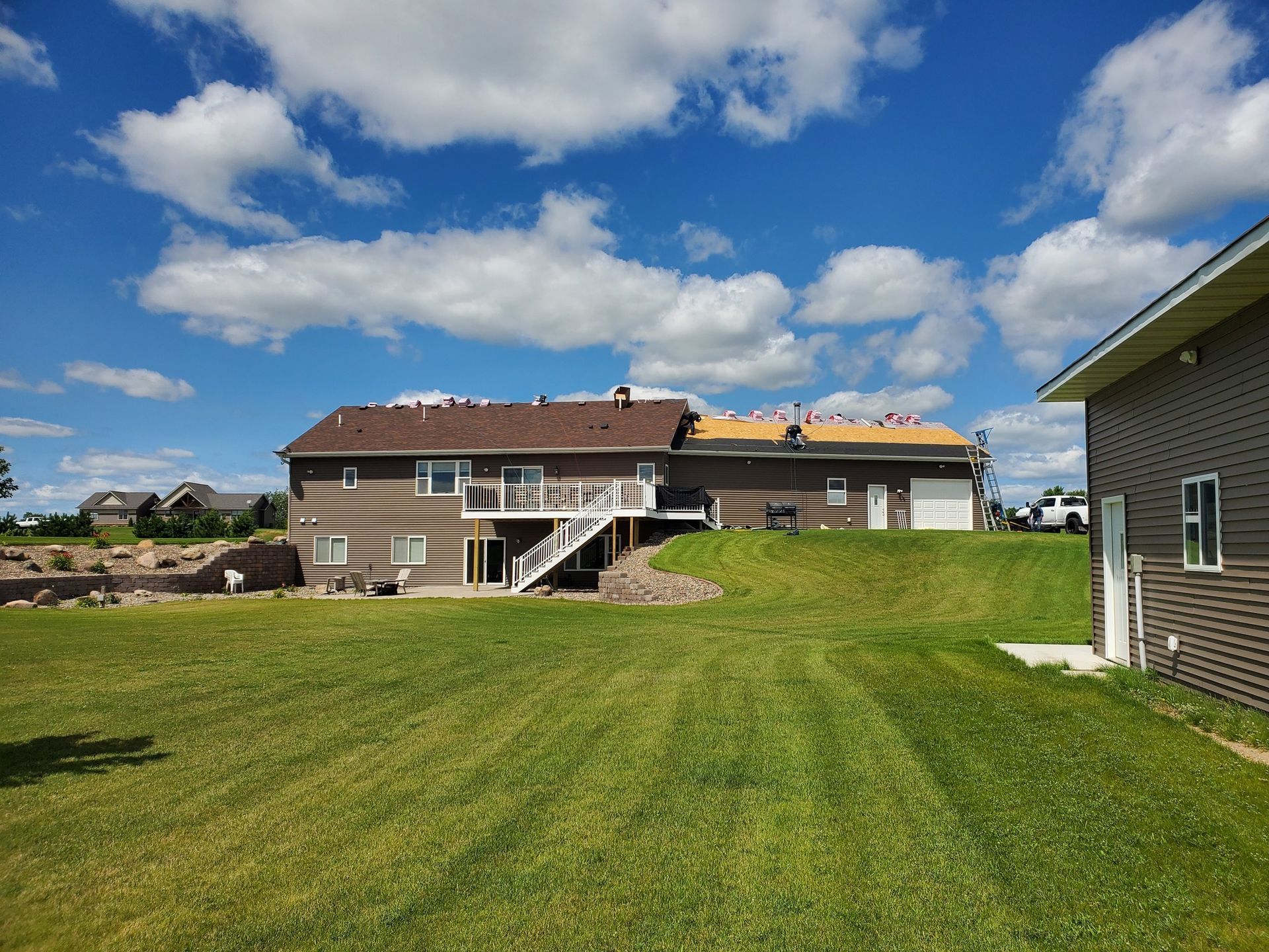 A brown house with a large backyard and deck under a blue sky with white clouds, showing ongoing roof work.