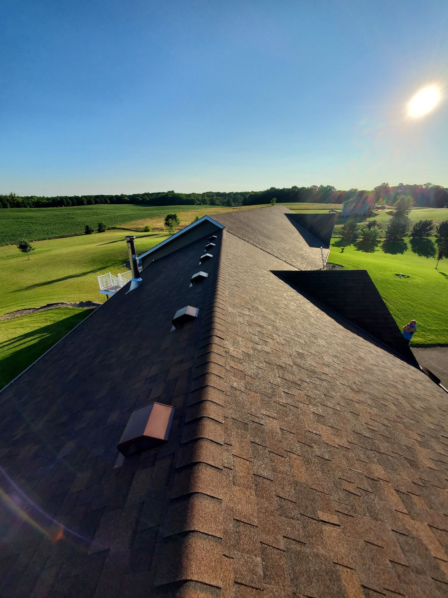 A high-angle view of a brown asphalt shingle roof, with several vents, overlooking a grassy field under a sunny sky.