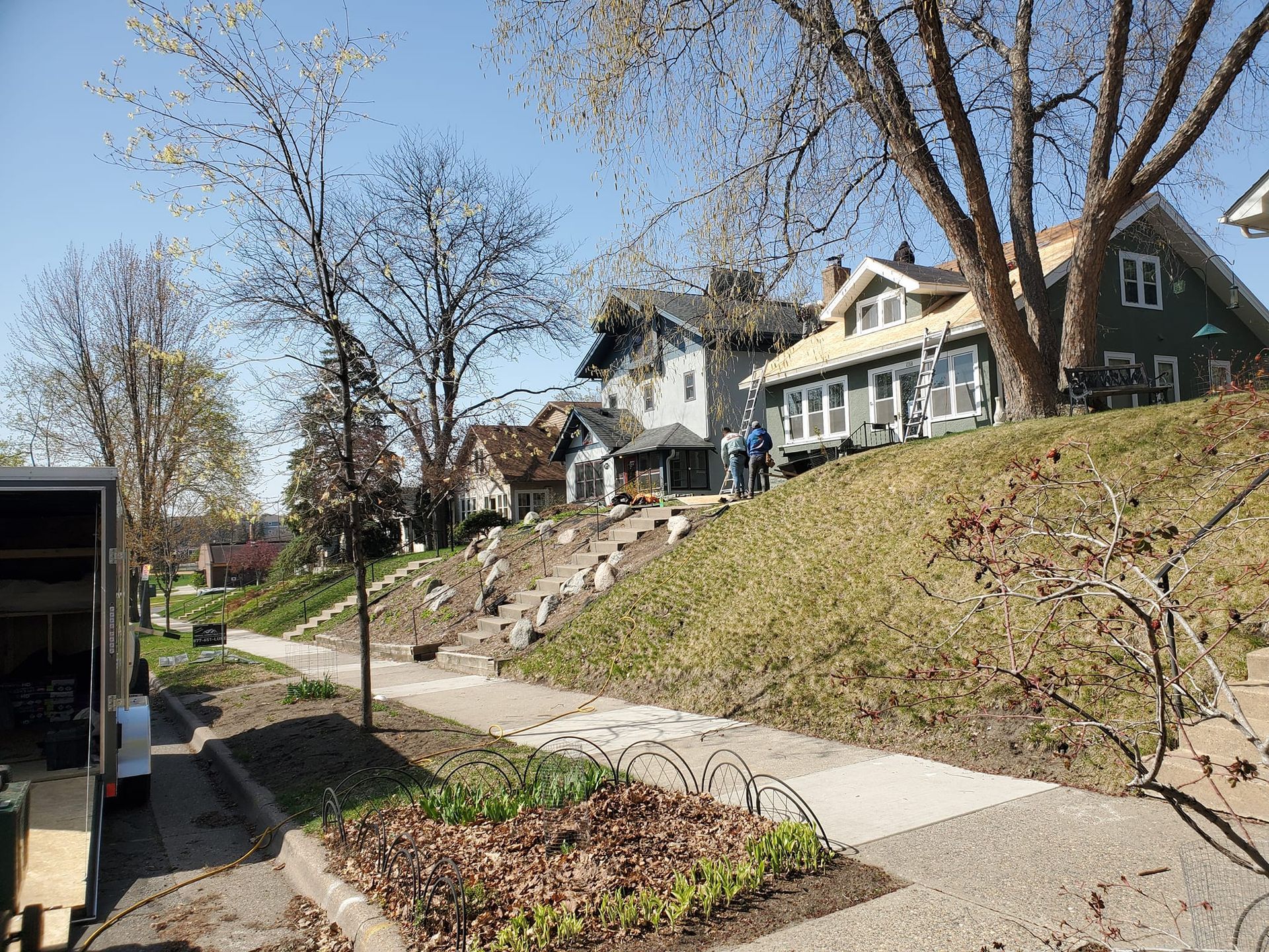 A residential street with houses on a grassy hillside, featuring stone steps, mature trees, and a parked trailer.