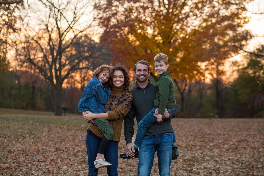 A family of four stands together in a field of autumn leaves at sunset, smiling at the camera.