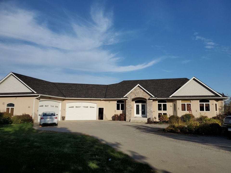 A large, beige, ranch-style house with a dark roof and a multi-car garage, viewed from a paved driveway under a blue sky.