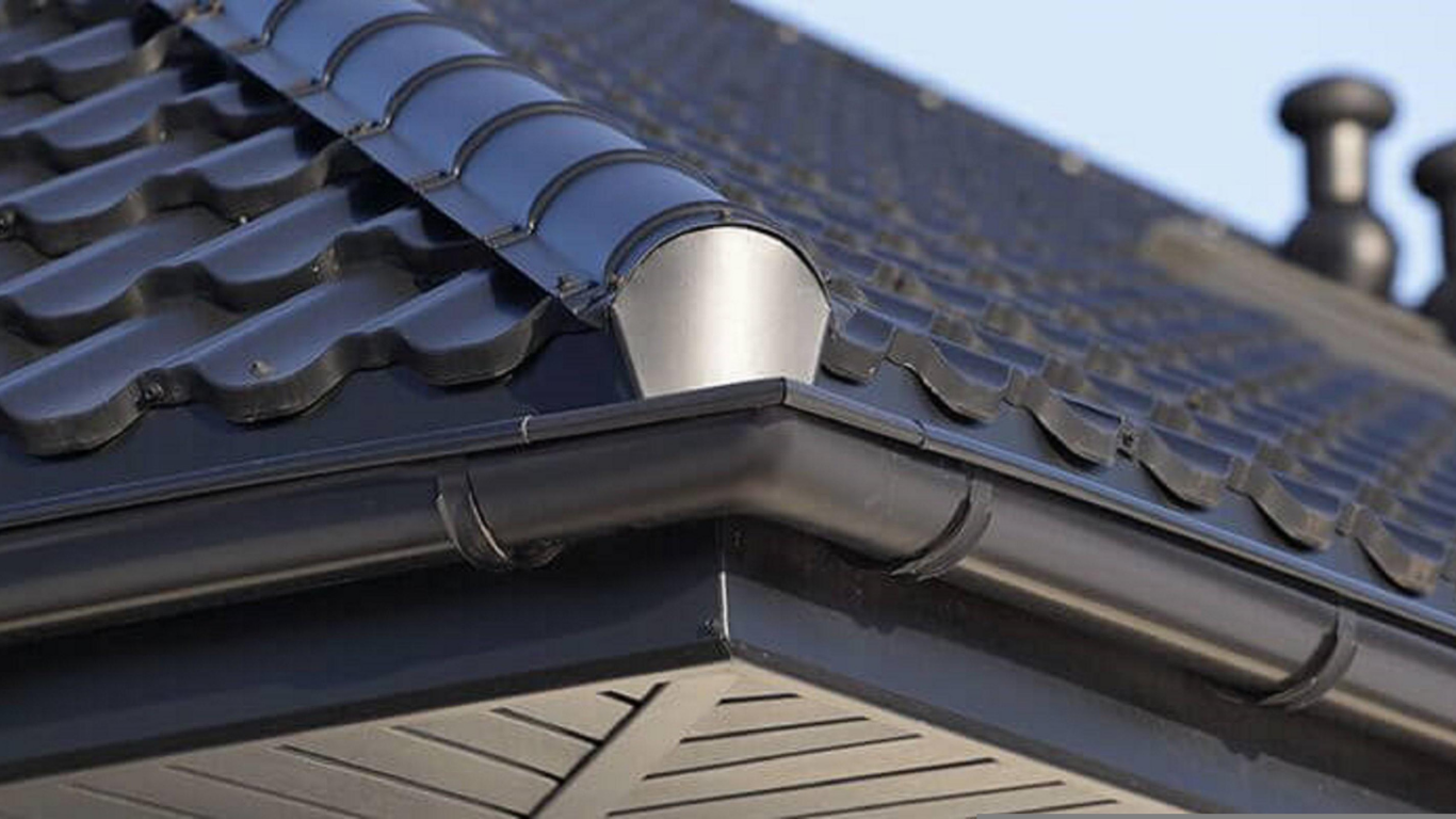 Close-up of a house roof with dark tiles and a white gutter under blue sky