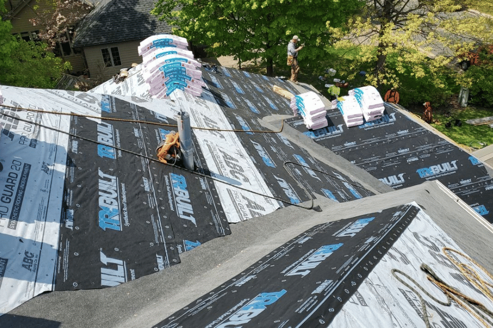 Workers installing black roof underlayment on a steep roof, with trees and a house in the background