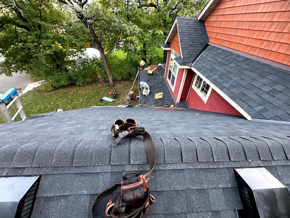 A tool belt rests on the dark shingled roof of a house under renovation, overlooking a yard and another roof section.