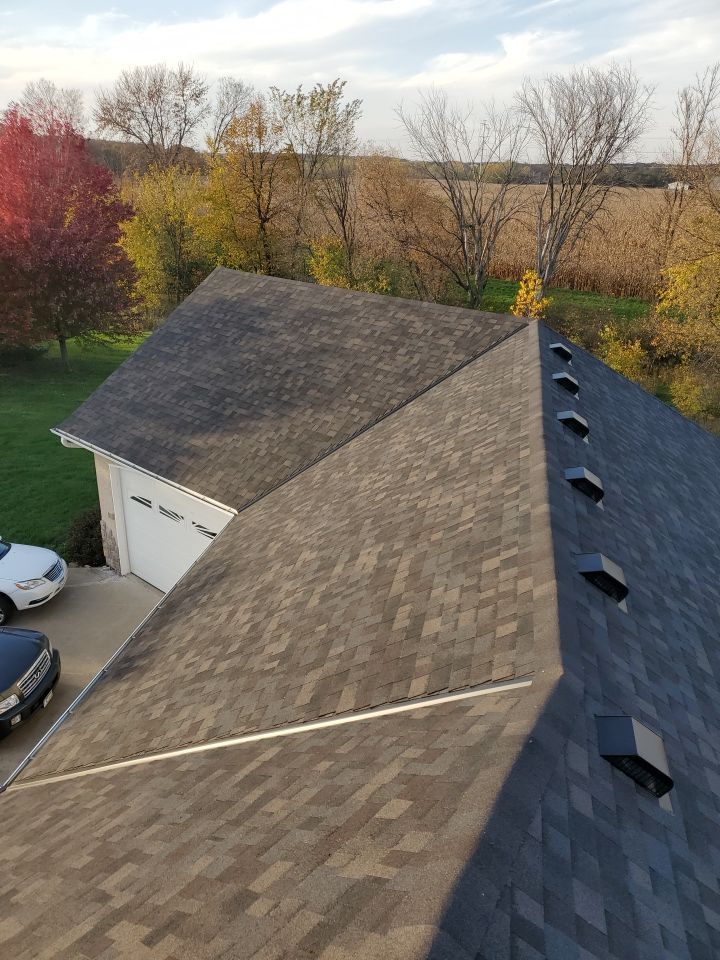 Aerial view of a shingled roof with skylights, beside a garage and parked cars, with autumn trees nearby