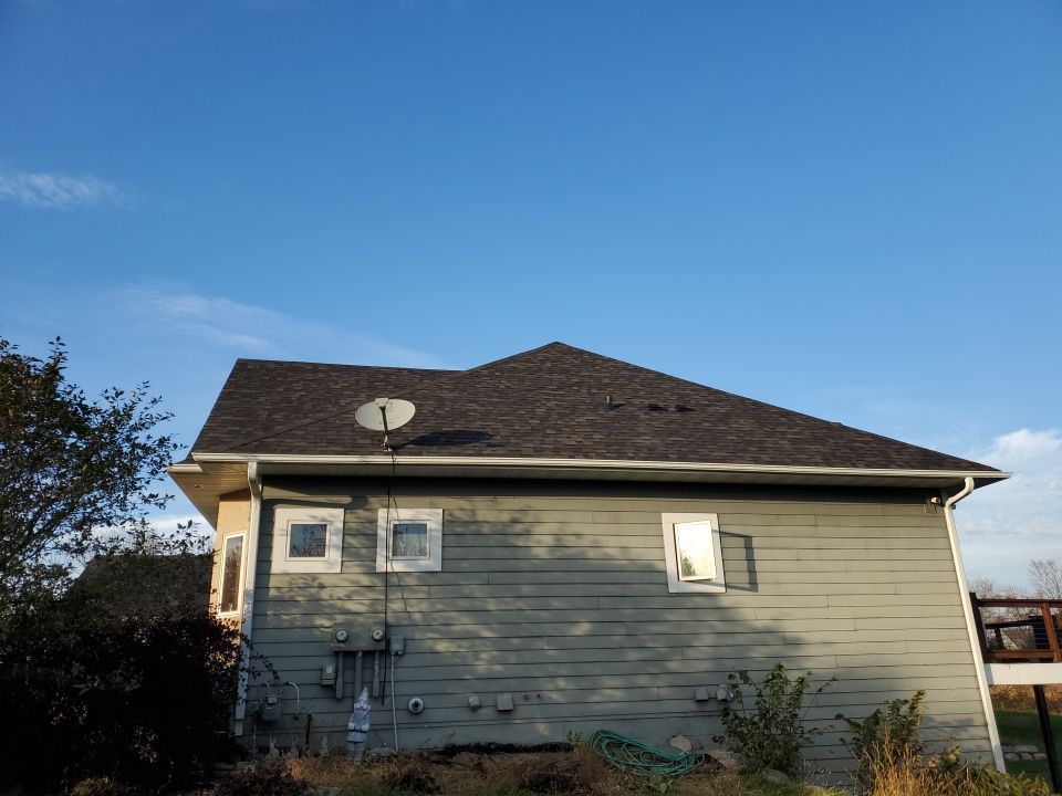 Back of a gray house with windows, utility meters, and a dark shingled roof under a blue sky