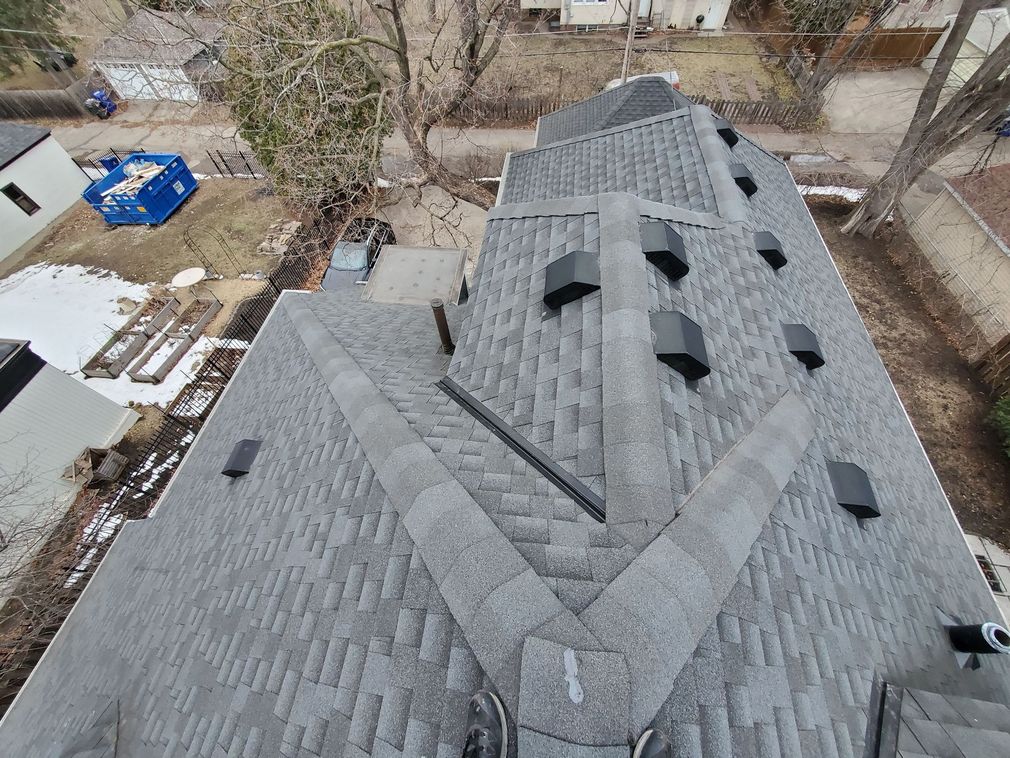 An aerial, high-angle view of a gray shingled residential roof with several dark vents, surrounded by a yard and trees.