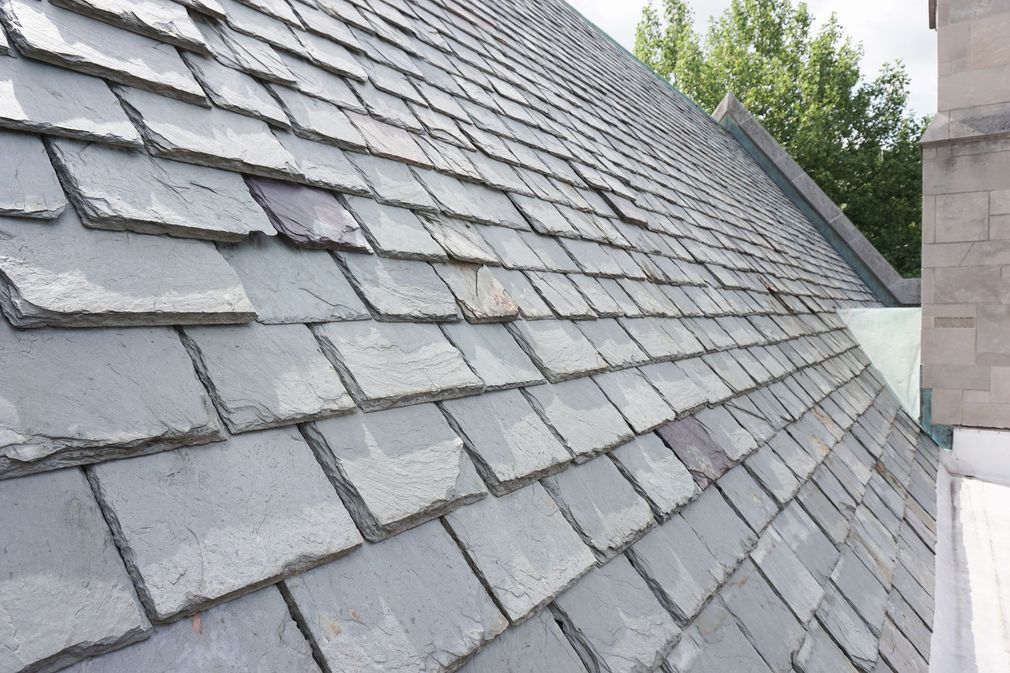 A sloped roof covered in gray asphalt shingles, with a wooden board resting on it and a security camera visible below.