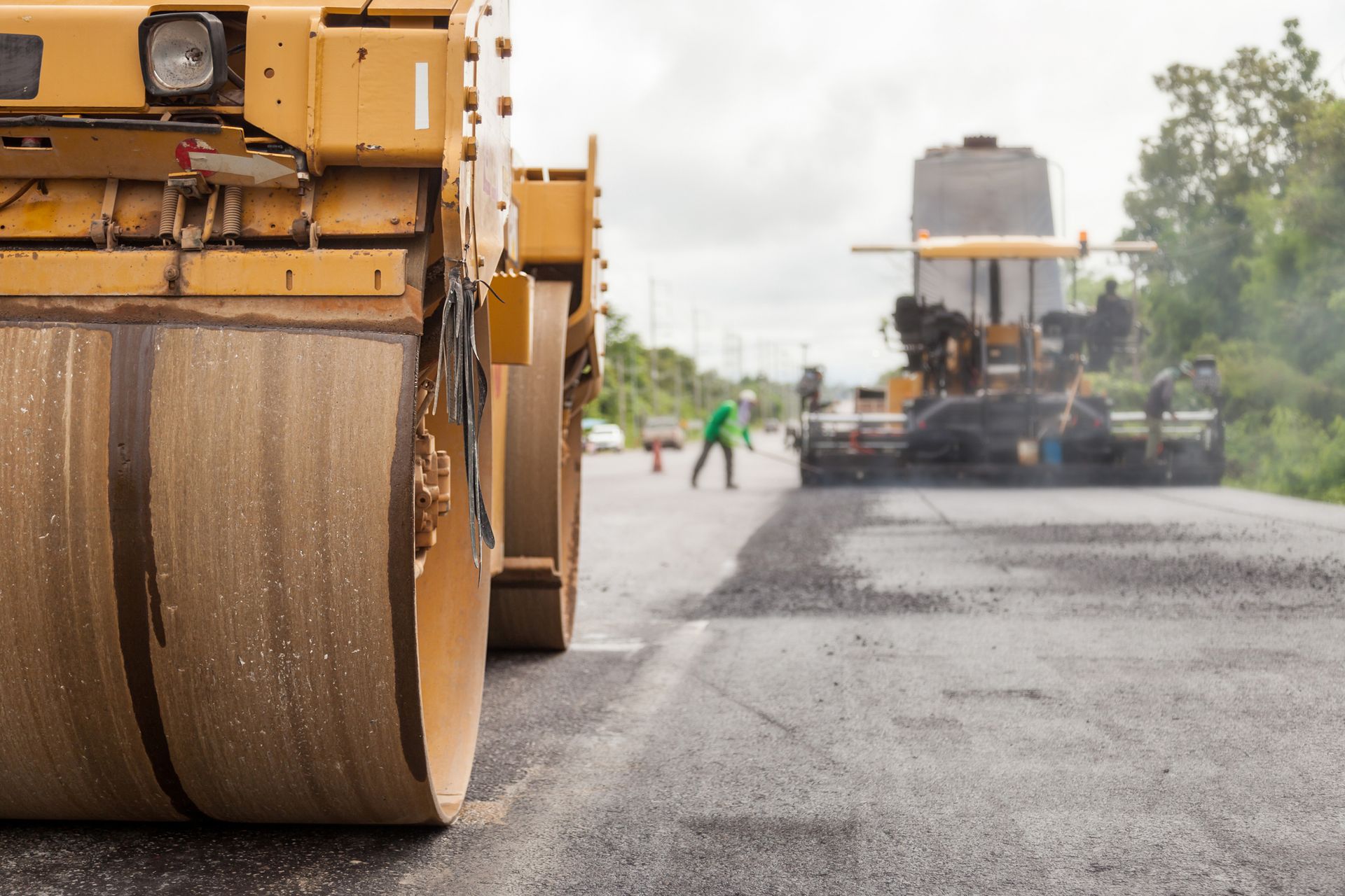 Yellow road roller compacting fresh asphalt on a road; paving equipment and worker in background.