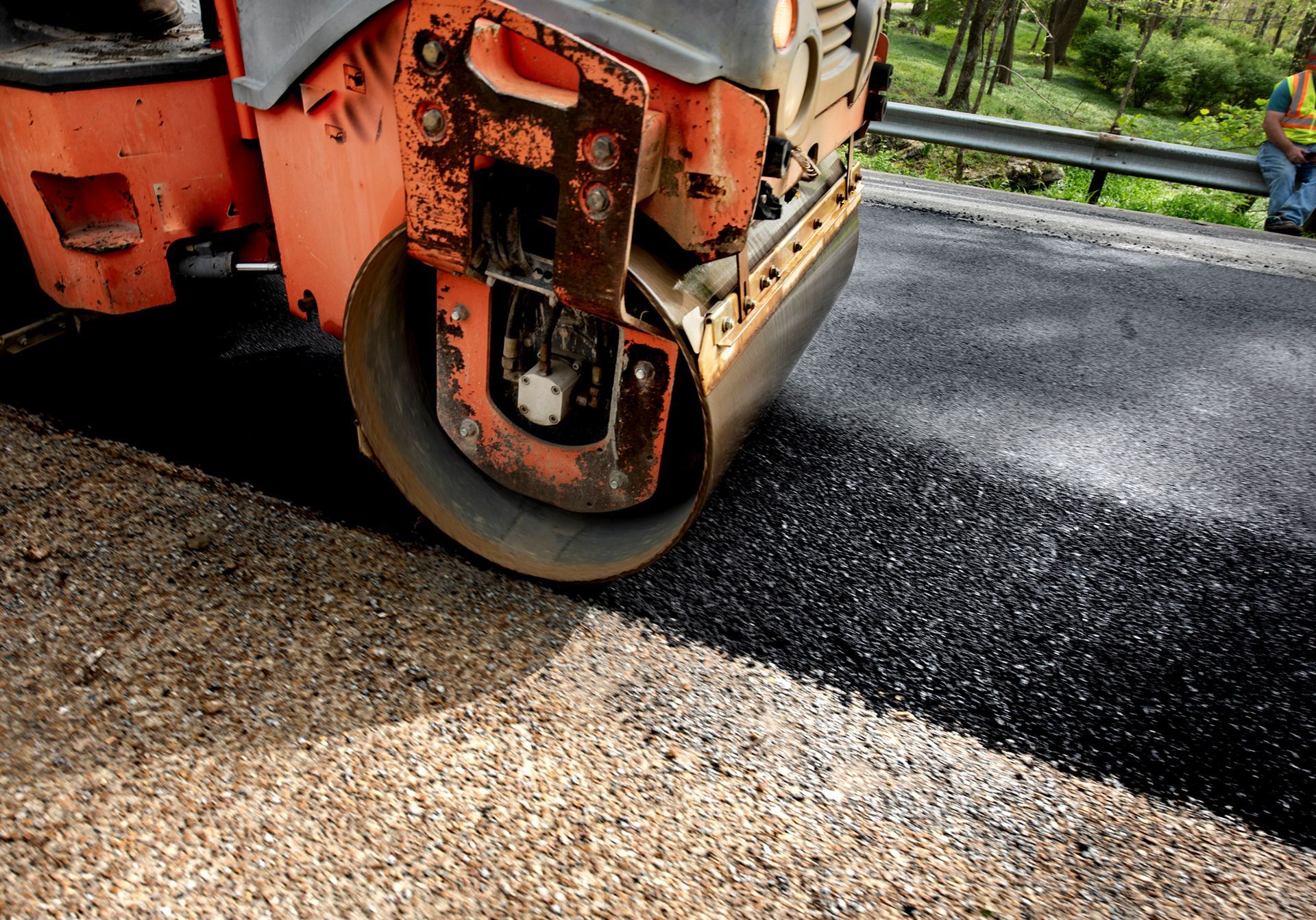 Road roller compacting fresh asphalt on a roadway; orange machine, black surface, rural setting. Road roller compacting fresh asphalt on a roadway; orange machine, black surface, rural setting.