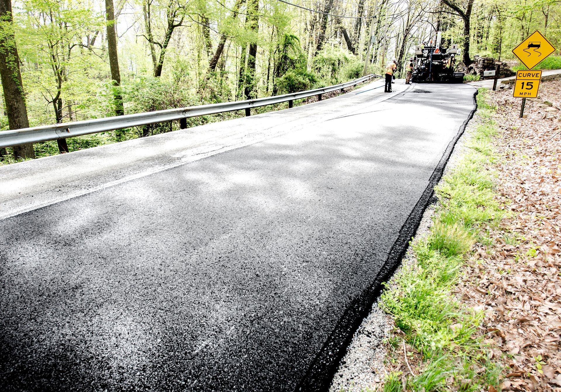 Freshly paved asphalt road next to a metal guardrail and grassy verge in a wooded area. Freshly paved asphalt road next to a metal guardrail and grassy verge in a wooded area.
