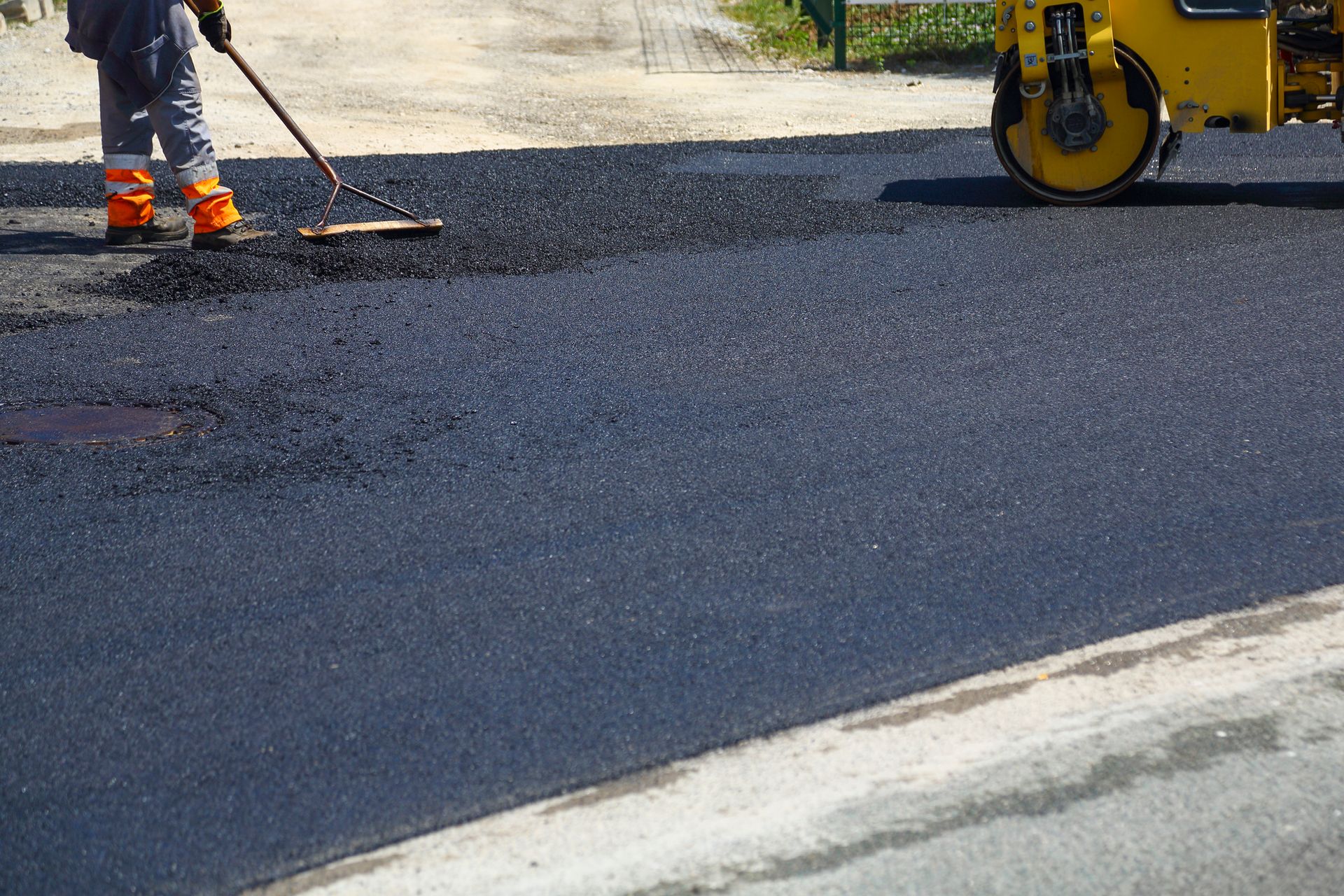 Worker using rake on fresh asphalt near a road roller.